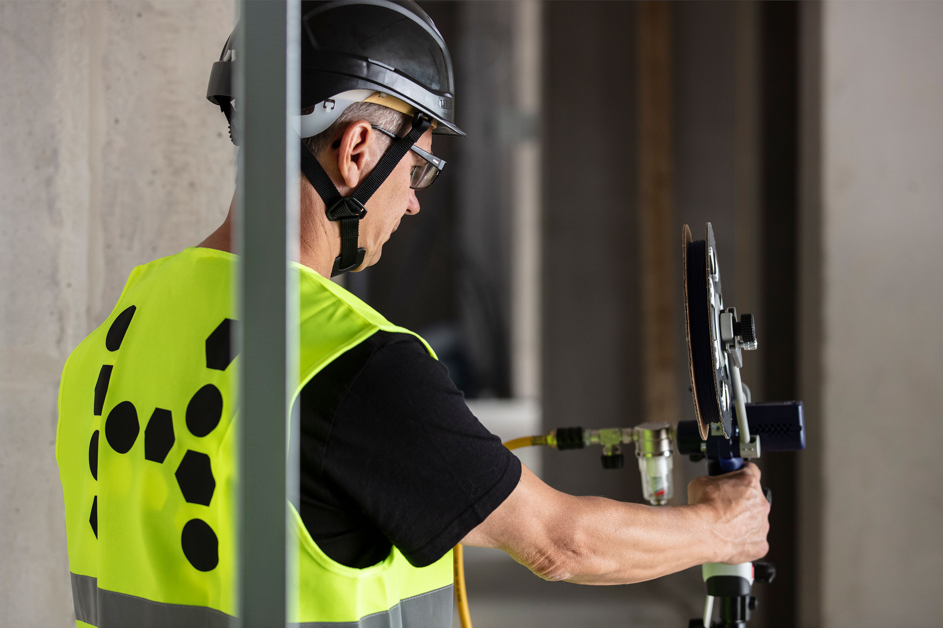 A person in a safety vest holds a Air Blown Fiber Installation Tool, ready to install optical fiber in an indoor construction area.