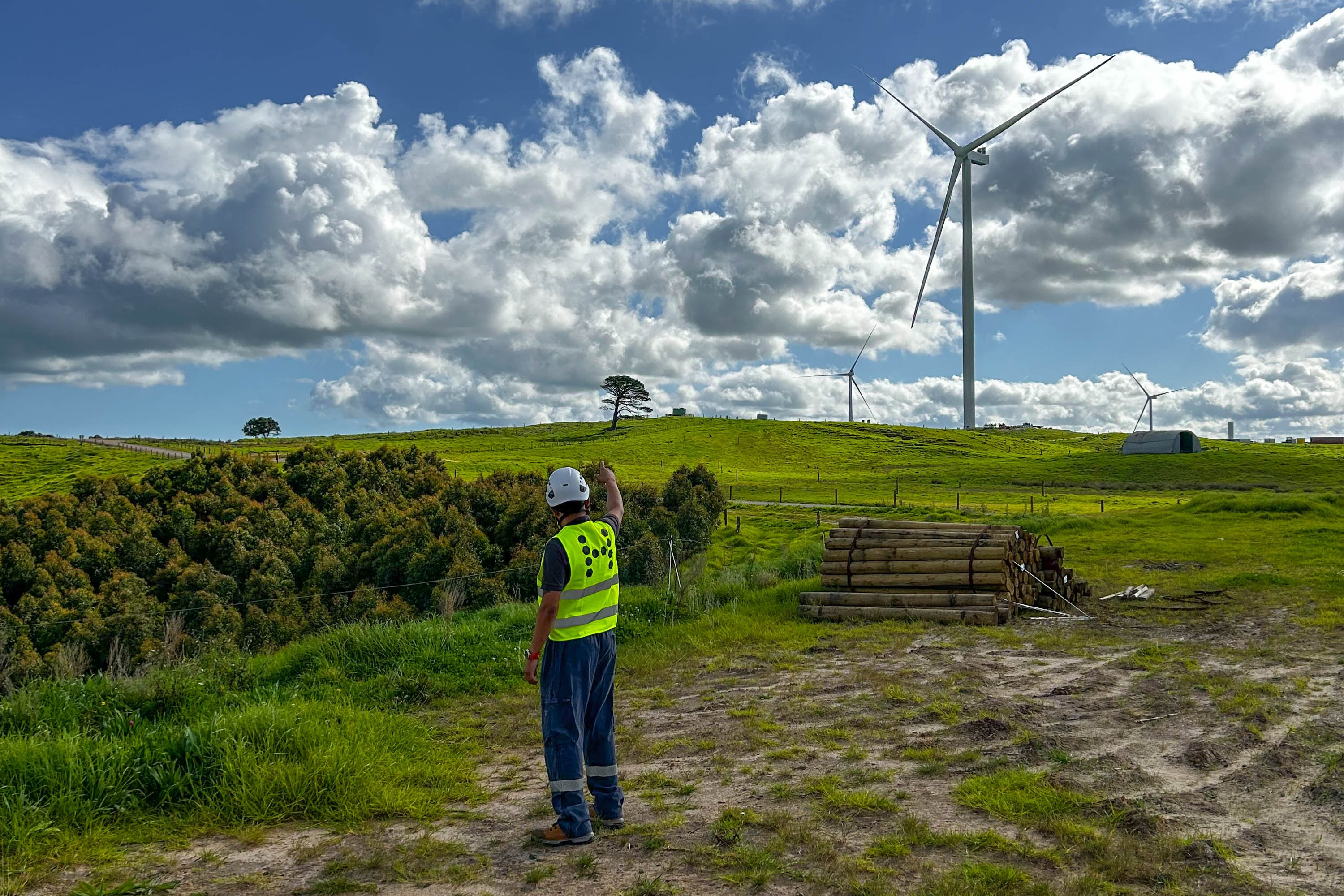 A person in a high-visibility vest is standing in an open field with wind mills on the horizon.